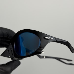 May include: Black sunglasses with blue lenses. The sunglasses have a sleek, modern design with a curved frame and the word "PRIZM" printed on the lens. The arms of the sunglasses are thin and curved.