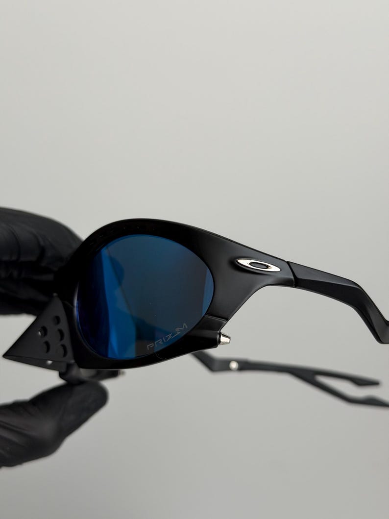 May include: Black sunglasses with blue lenses. The sunglasses have a sleek, modern design with a curved frame and the word "PRIZM" printed on the lens. The arms of the sunglasses are thin and curved.
