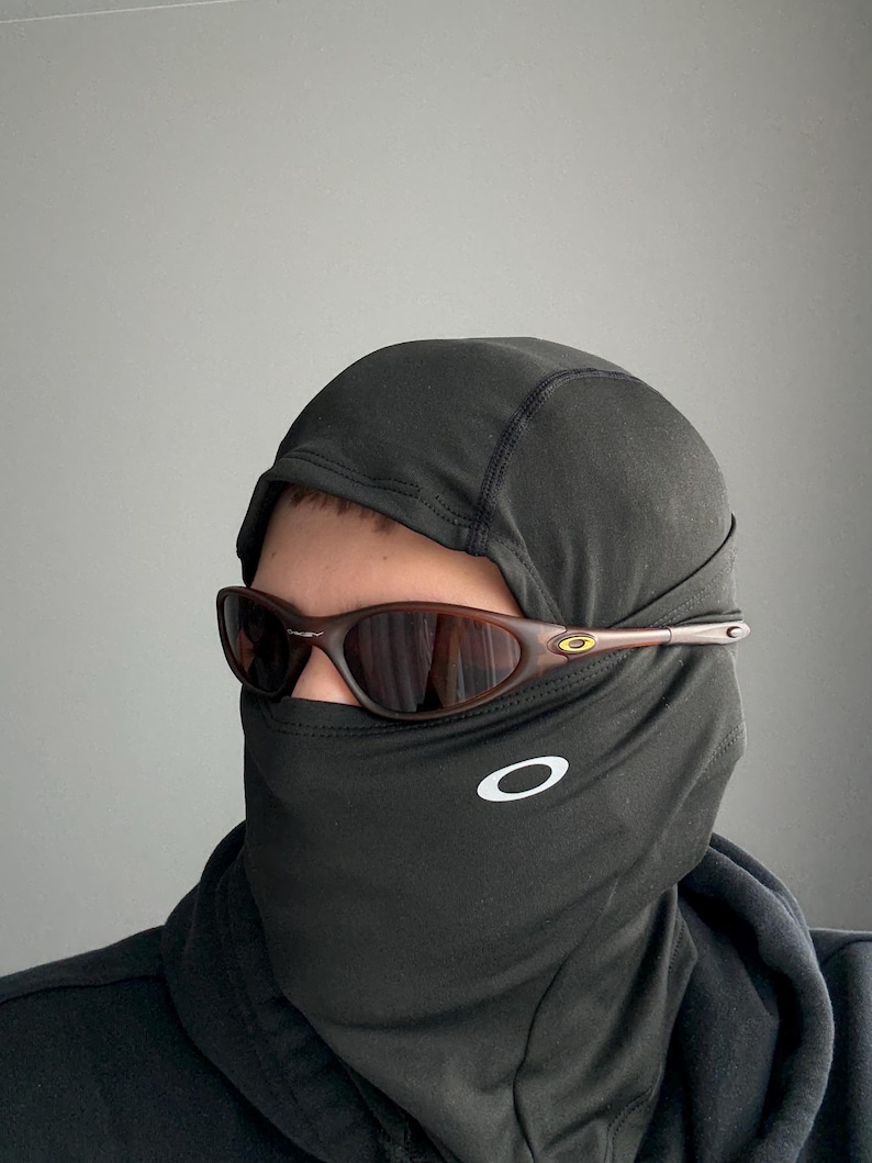 May include: Brown-framed sunglasses with a dark lens are worn over a black balaclava. The balaclava has a white Oakley logo on the front. The sunglasses have a small gold accent near the temple.