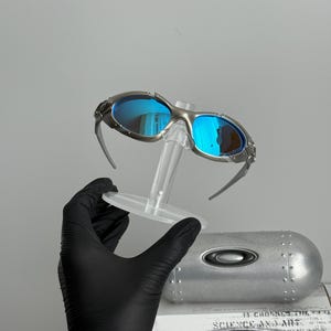 May include: Silver-framed sunglasses with blue mirrored lenses. The sunglasses are displayed on a clear stand, with a silver case and a box with the text "SCIENCE AND ART" in the background.