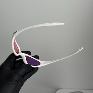 May include: White sunglasses with purple and pink lenses. The sunglasses have a sleek, modern design with a curved frame and a small logo on the temple. The lenses are a gradient color, transitioning from pink to purple.