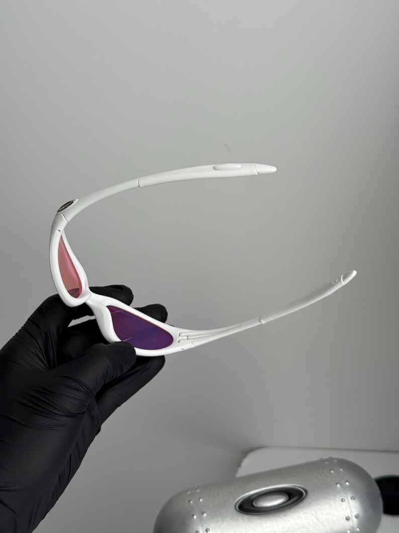 May include: White sunglasses with purple and pink lenses. The sunglasses have a sleek, modern design with a curved frame and a small logo on the temple. The lenses are a gradient color, transitioning from pink to purple.