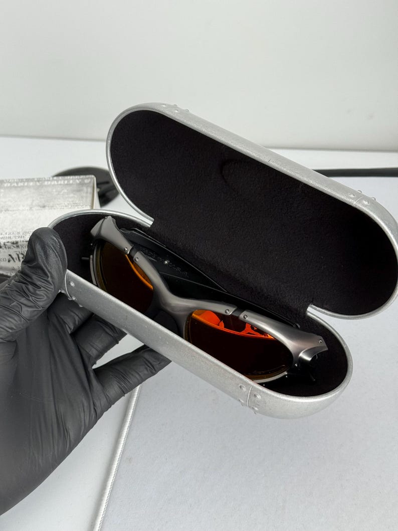 May include: A pair of sunglasses with orange lenses rests inside an open, silver-colored case. The sunglasses have a dark frame. The case is lined with black fabric.