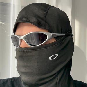 Puede incluir: Una balaclava negra con el logotipo blanco de Oakley en la parte delantera, combinada con gafas de sol con montura plateada y lentes oscuras. La balaclava cubre la cabeza y el cuello.
