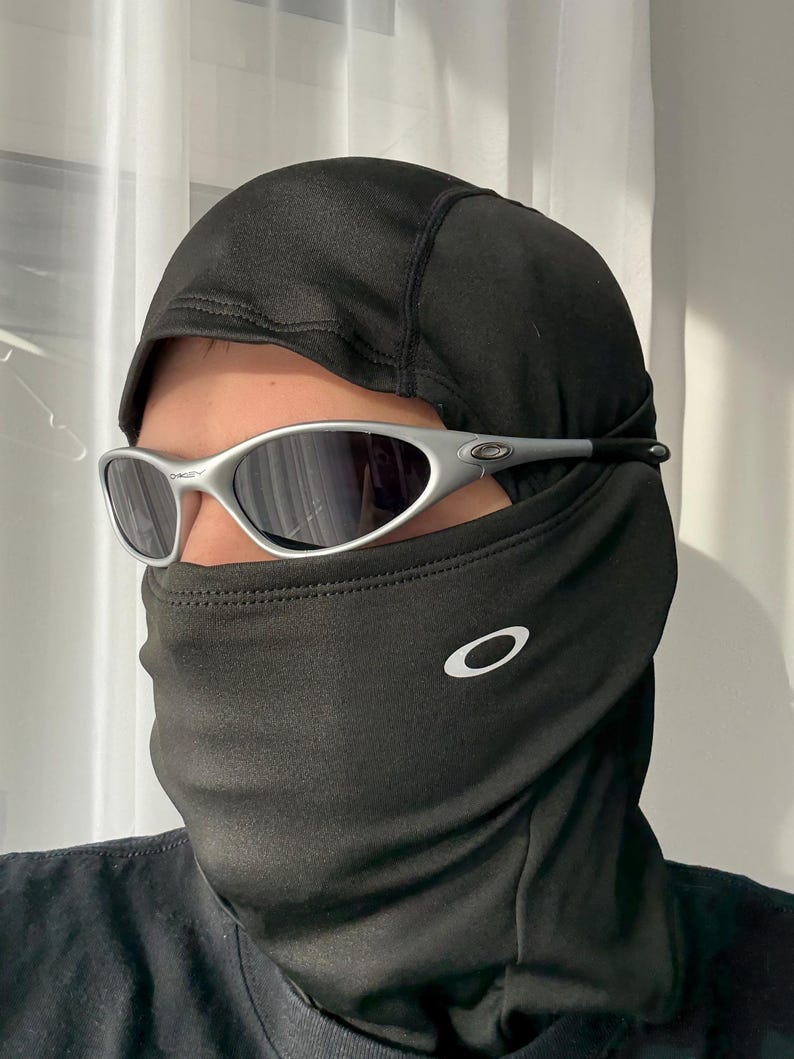 Puede incluir: Una balaclava negra con el logotipo blanco de Oakley en la parte delantera, combinada con gafas de sol con montura plateada y lentes oscuras. La balaclava cubre la cabeza y el cuello.