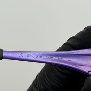 May include: Close-up of a purple object with the word "jacket" etched into the surface. The object is held by a gloved hand against a white background. The object has a black tip.