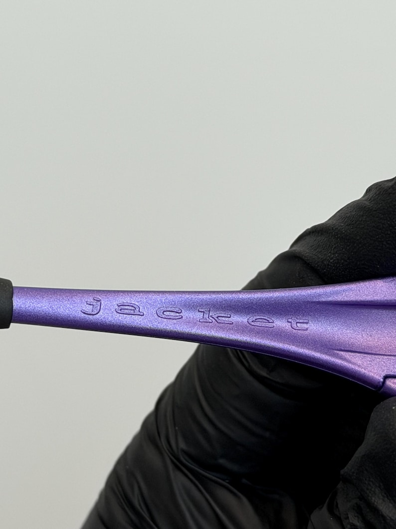 May include: Close-up of a purple object with the word "jacket" etched into the surface. The object is held by a gloved hand against a white background. The object has a black tip.