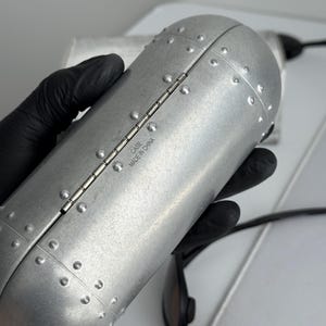 Puede incluir: Un estuche cil&iacute;ndrico plateado para gafas de sol con un dise&ntilde;o remachado. El estuche est&aacute; sujeto por una mano enguantada y tiene el texto "CASE MADE IN CHINA" impreso en el lateral. Se ven gafas de sol negras.