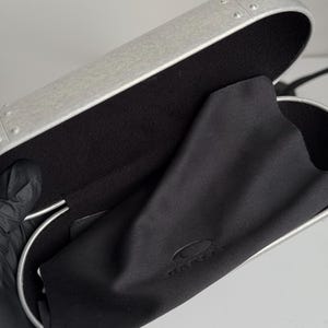 May include: A silver-colored glasses case with a black interior and a black microfiber cleaning cloth. The case has a riveted design and a curved shape. The cloth has the brand name printed on it.