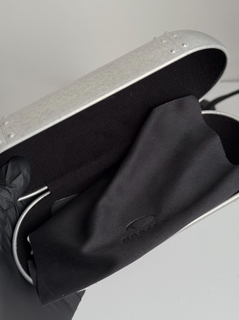 May include: A silver-colored glasses case with a black interior and a black microfiber cleaning cloth. The case has a riveted design and a curved shape. The cloth has the brand name printed on it.