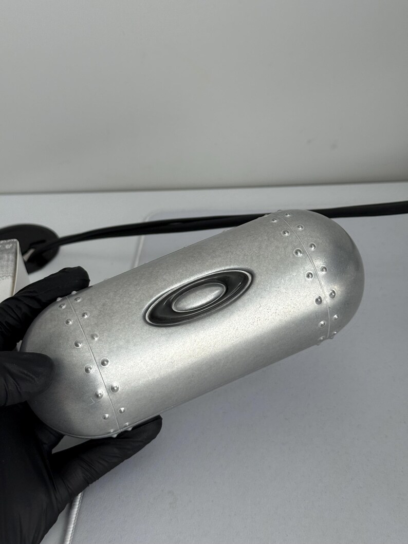 May include: Silver-colored eyewear case with a riveted design. The case has a rounded shape and features the Oakley logo in black. The case is held by a gloved hand.
