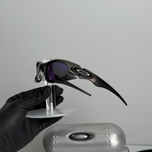 May include: Black and silver sunglasses with purple lenses are displayed on a clear stand. A silver case with the Oakley logo is below the sunglasses. The text "SCIENCE AND ART" is visible on the box.