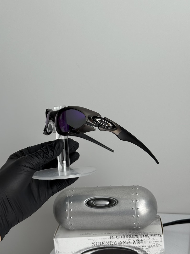 May include: Black and silver sunglasses with purple lenses are displayed on a clear stand. A silver case with the Oakley logo is below the sunglasses. The text "SCIENCE AND ART" is visible on the box.