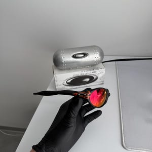 May include: Black sunglasses with red mirrored lenses and a flame design on the temples. The sunglasses are held by a gloved hand. A silver case and a box with the Oakley logo are in the background.