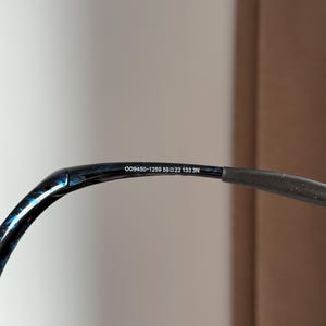 Puede incluir: Primer plano de la patilla de unas gafas. La montura es negra con un patr&oacute;n veteado azul y negro. El texto "OO9450-1259 59 22 133 3N" est&aacute; impreso en la patilla.