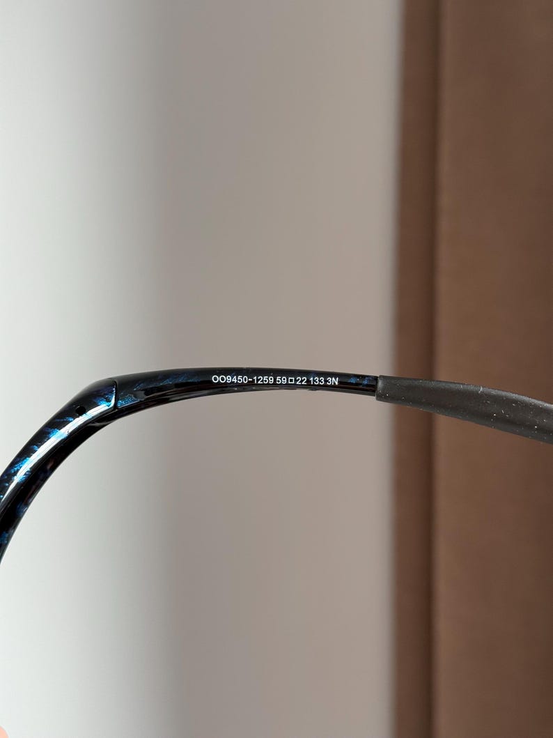 Puede incluir: Primer plano de la patilla de unas gafas. La montura es negra con un patr&oacute;n veteado azul y negro. El texto "OO9450-1259 59 22 133 3N" est&aacute; impreso en la patilla.