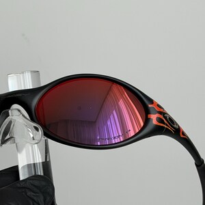 May include: Black sunglasses with red lenses and orange flame detailing on the temples. The lenses reflect a purple hue. The sunglasses are held up by a clear stand.