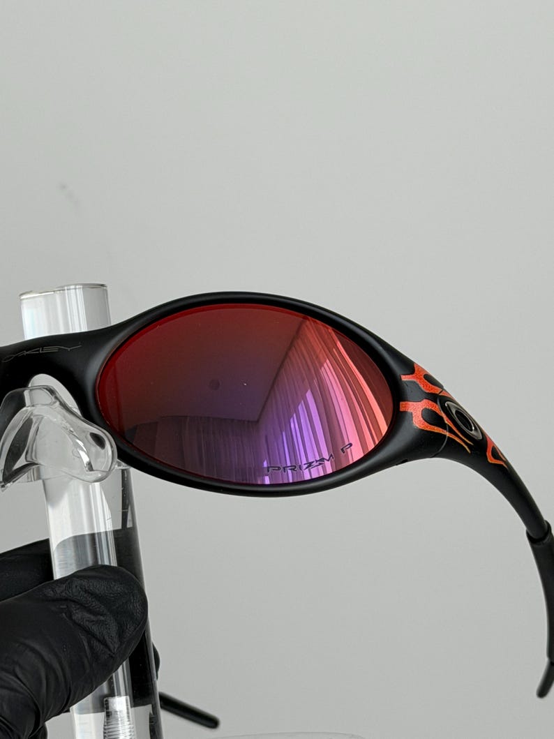 May include: Black sunglasses with red lenses and orange flame detailing on the temples. The lenses reflect a purple hue. The sunglasses are held up by a clear stand.