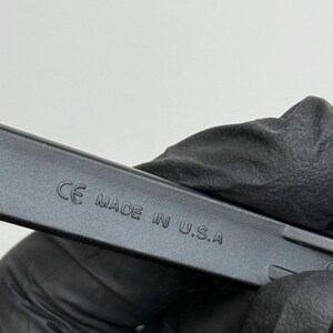May include: Close-up of a black object with the text "CE MADE IN U.S.A." embossed on its surface. The object is held by a gloved hand against a plain white background.