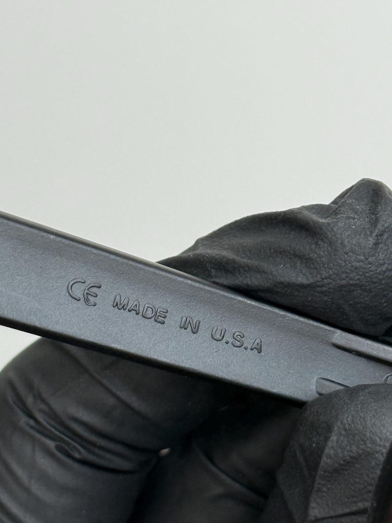 May include: Close-up of a black object with the text "CE MADE IN U.S.A." embossed on its surface. The object is held by a gloved hand against a plain white background.