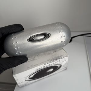 May include: A silver-colored eyewear case with a riveted design and the Oakley logo. The case is resting on a white box with the words "SCIENCE AND ART" printed on it.