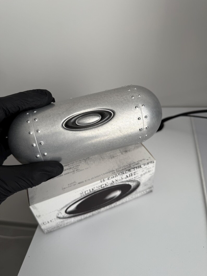 May include: A silver-colored eyewear case with a riveted design and the Oakley logo. The case is resting on a white box with the words "SCIENCE AND ART" printed on it.