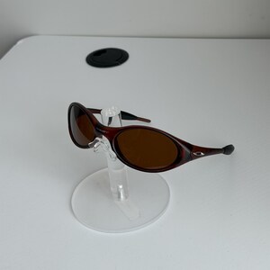 May include: Brown oval sunglasses with a dark frame, displayed on a clear stand. The lenses are a warm brown color. The sunglasses have a sleek, modern design.