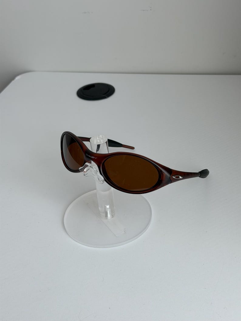 May include: Brown oval sunglasses with a dark frame, displayed on a clear stand. The lenses are a warm brown color. The sunglasses have a sleek, modern design.
