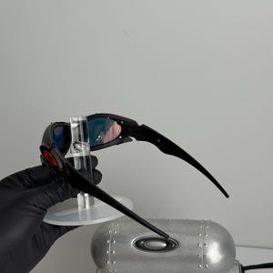 May include: Black and red sunglasses with a unique design. The lenses have a gradient of colors, and the frame is black. The sunglasses are held up by a clear stand. A silver case with the Oakley logo is in the background.