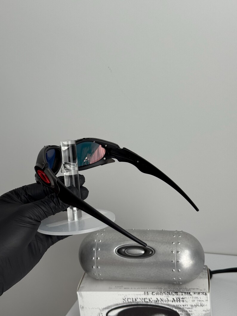 May include: Black and red sunglasses with a unique design. The lenses have a gradient of colors, and the frame is black. The sunglasses are held up by a clear stand. A silver case with the Oakley logo is in the background.