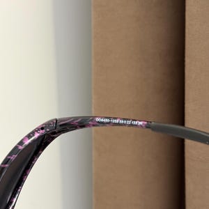 May include: Close-up of sunglasses with a black and pink patterned frame. The text "OO9450-1259 59 22 133" is printed on the frame. The lenses are dark, and the arms are a dark gray color.