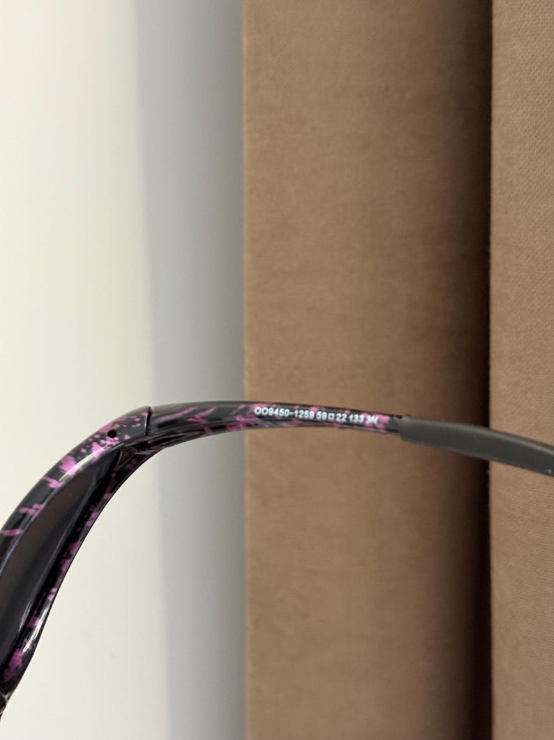 May include: Close-up of sunglasses with a black and pink patterned frame. The text "OO9450-1259 59 22 133" is printed on the frame. The lenses are dark, and the arms are a dark gray color.