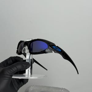 May include: Black sunglasses with blue lenses are displayed on a clear stand. The sunglasses have a sleek, modern design with a blue accent on the frame. A silver case with the brand logo is visible below.