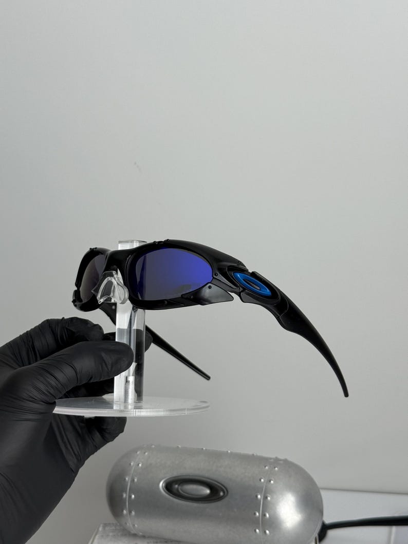 May include: Black sunglasses with blue lenses are displayed on a clear stand. The sunglasses have a sleek, modern design with a blue accent on the frame. A silver case with the brand logo is visible below.