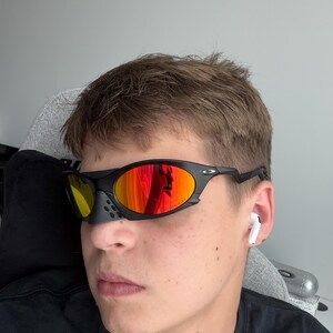 May include: Black sports sunglasses with orange mirrored lenses. The sunglasses have a black frame and a small nose piece. The lenses reflect the surrounding environment. The person is wearing a black shirt and has white earbuds in their ears.