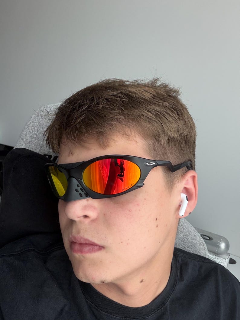 May include: Black sports sunglasses with orange mirrored lenses. The sunglasses have a black frame and a small nose piece. The lenses reflect the surrounding environment. The person is wearing a black shirt and has white earbuds in their ears.