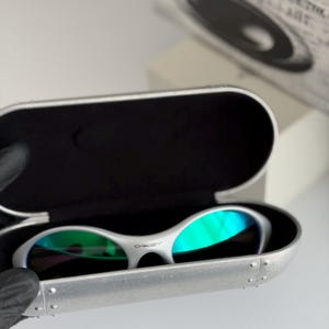May include: White Oakley sunglasses with green mirrored lenses sit in an open silver case. The case has a black interior and is riveted along the edges. The Oakley logo is visible on the sunglasses.