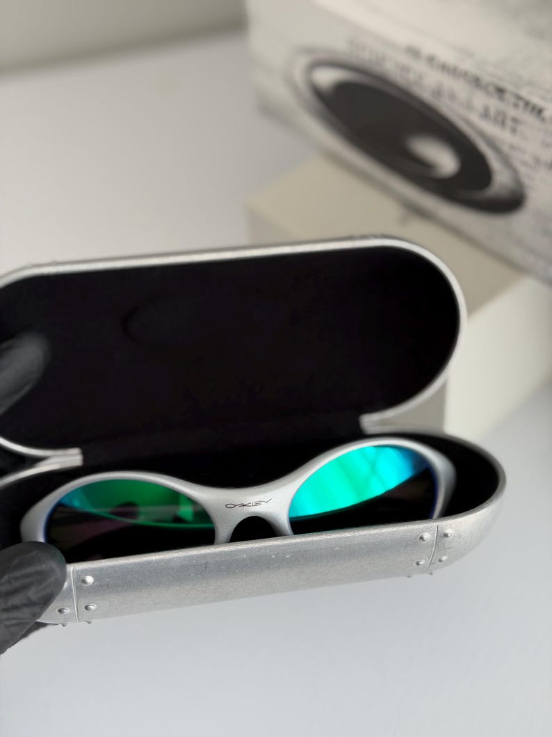 May include: White Oakley sunglasses with green mirrored lenses sit in an open silver case. The case has a black interior and is riveted along the edges. The Oakley logo is visible on the sunglasses.