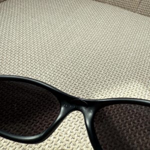 May include: Black sunglasses with dark lenses. The frame is a matte black, and the word "MINISTE" is printed on the bridge. The sunglasses are resting on a textured, light-coloured surface.