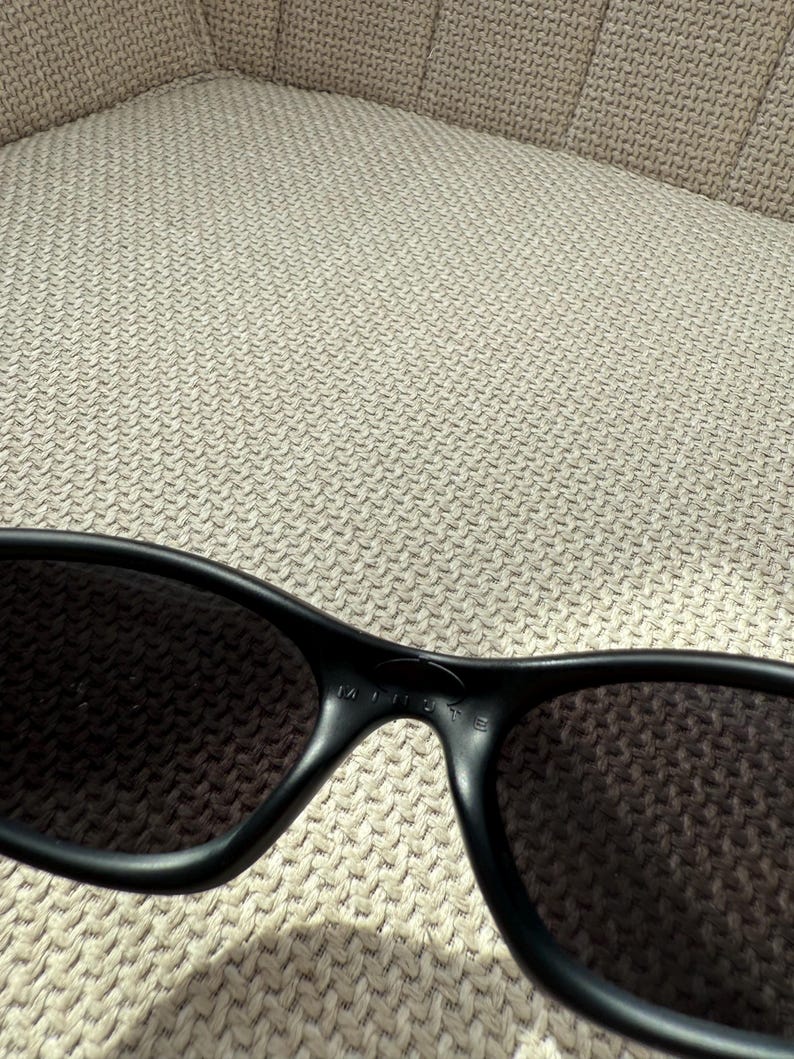 May include: Black sunglasses with dark lenses. The frame is a matte black, and the word "MINISTE" is printed on the bridge. The sunglasses are resting on a textured, light-coloured surface.