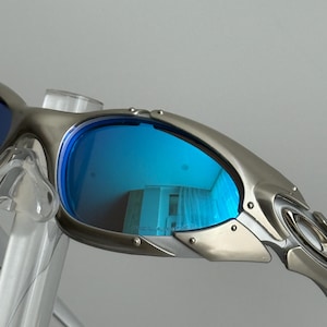 May include: Close-up of silver-framed sunglasses with blue lenses. The lenses reflect a room with curtains. The sunglasses have a sleek, modern design with visible rivets and a curved temple.