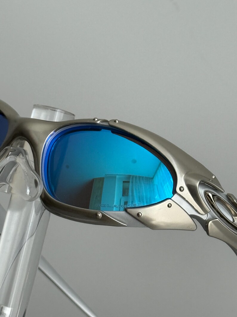 May include: Close-up of silver-framed sunglasses with blue lenses. The lenses reflect a room with curtains. The sunglasses have a sleek, modern design with visible rivets and a curved temple.