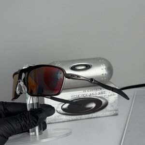 May include: Black and silver-framed sunglasses with red-tinted lenses. The sunglasses are displayed on a clear stand, with a silver case and a box with the text "SCIENCE AND ART" in the background.