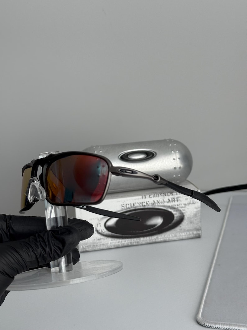 May include: Black and silver-framed sunglasses with red-tinted lenses. The sunglasses are displayed on a clear stand, with a silver case and a box with the text "SCIENCE AND ART" in the background.