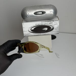 May include: Olive green and brown sunglasses with a matching case and box. The sunglasses have brown lenses and a unique frame design. The case is silver and cylindrical, with the brand logo. The box has the text "SCIENCE AND ART".