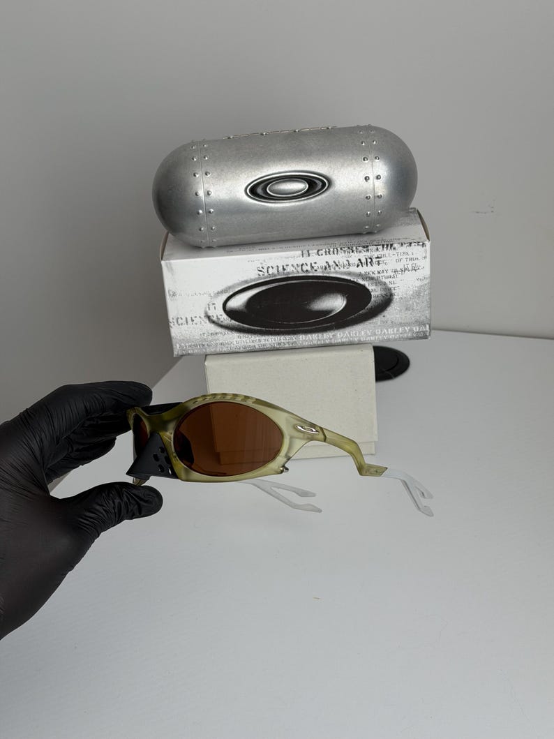 May include: Olive green and brown sunglasses with a matching case and box. The sunglasses have brown lenses and a unique frame design. The case is silver and cylindrical, with the brand logo. The box has the text "SCIENCE AND ART".