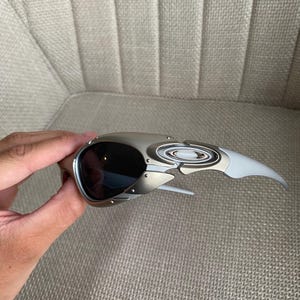 May include: Silver and black sunglasses with a unique, angular design. The sunglasses have dark lenses and a metallic frame with a white arm. The glasses are held by a hand.