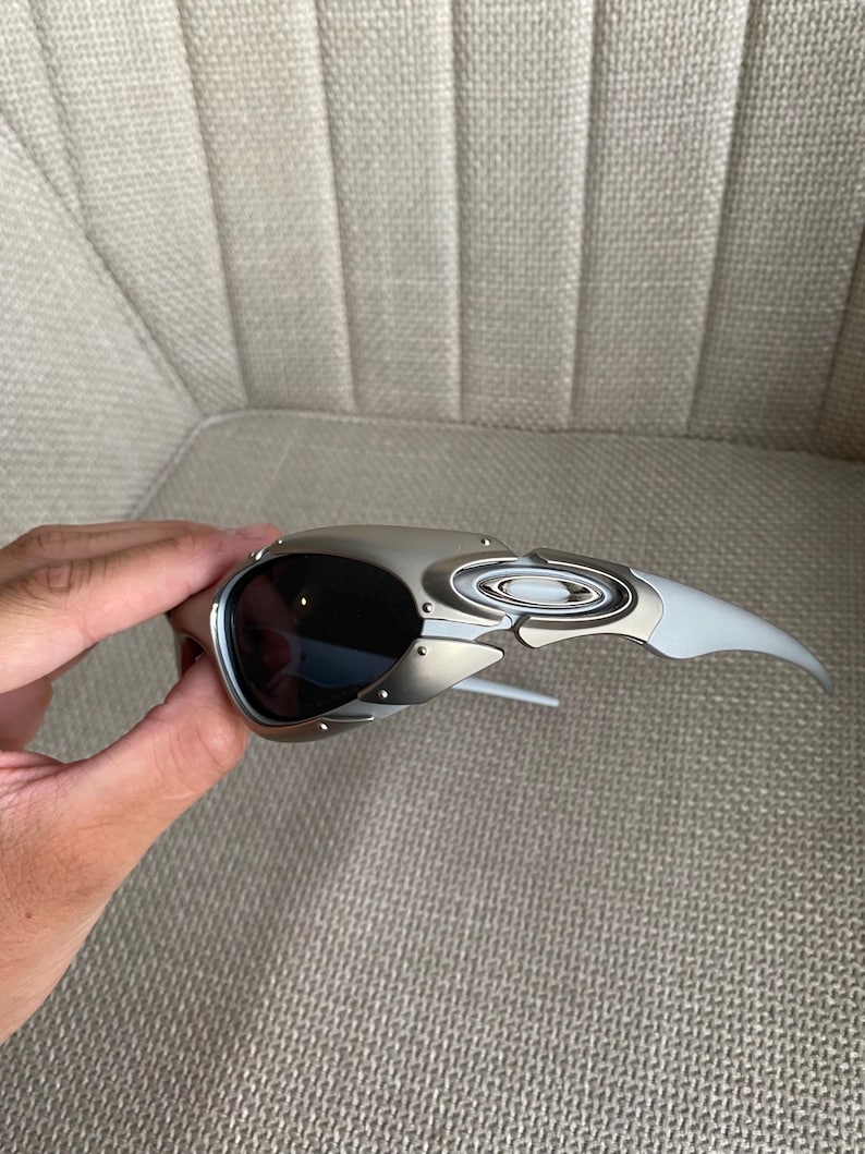 May include: Silver and black sunglasses with a unique, angular design. The sunglasses have dark lenses and a metallic frame with a white arm. The glasses are held by a hand.