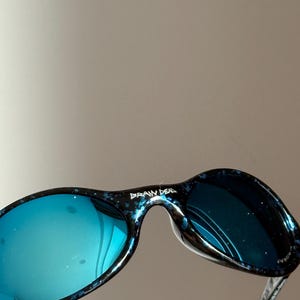 May include: Sunglasses with blue-tinted lenses and a speckled black and blue frame. The text "BRAIN DEAD" is printed on the top of the frame. The lenses reflect the surroundings. The design is sleek and modern.
