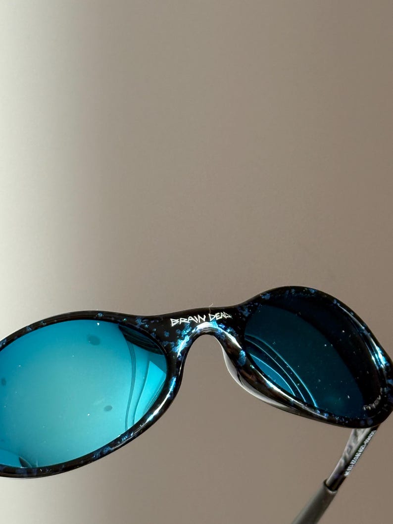May include: Sunglasses with blue-tinted lenses and a speckled black and blue frame. The text "BRAIN DEAD" is printed on the top of the frame. The lenses reflect the surroundings. The design is sleek and modern.